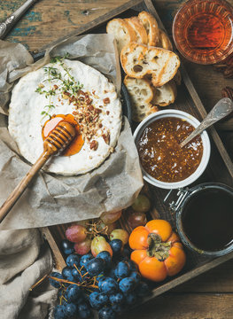 Cheese, fruit and wine set. Camembert in small pan with nuts and herbs, grapes, persimmon, fig jam, honey, baguette slices and glass of rose wine over rustic wooden background, top view