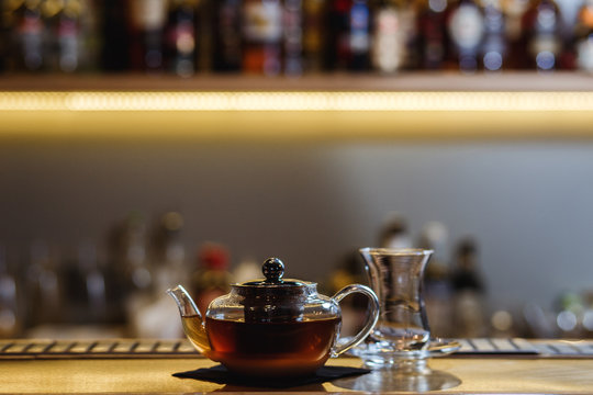 An underexposed lifestyle horizontal image of a teapot with hot alcoholic cocktail on a black tea basis. Wooden bar stand of a restaurant.