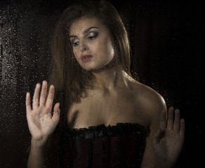 Portrait of a sensual young sexy woman wearing corset, touching her hair and posing behind transparent glass covered by water drops.