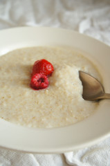 Oatmeal porridge with two red berries in white round plate on the white tablecloth vertical