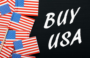 The words Buy USA in white text on a blackboard next to United States of America flags as a reminder to buy American products and services