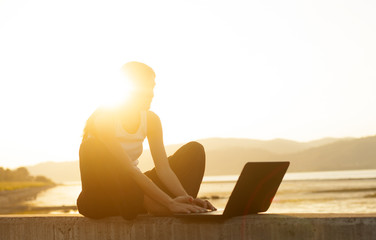 Young woman by the river in sunset with laptop