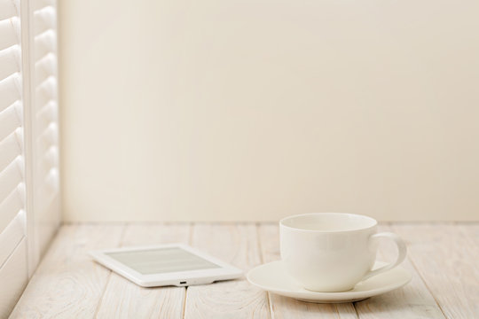 E-book and a cup on a light wooden background near a window with