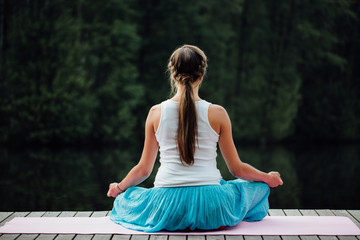 Young woman in the lotus position is practicing yoga in the forest next to the river. sitting on mats  the wooden pier.