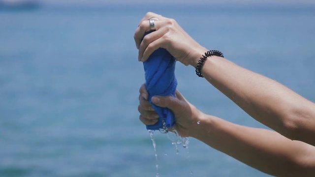 Close up of hands of young woman squeezing blue towel after swimming on the beach of Bali island. Female traveler with bracelet and ring holding terry-cloth and pressing it with sea in the background.