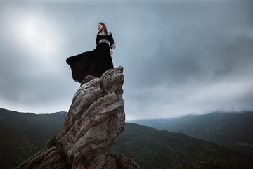 Young woman in long dress enjoying nature on the mountains. The girl in black dress with long flying train stands on the top of the rock - 132265353