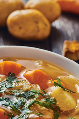 Vegetable soup in white bowl on wooden table potatoes in background