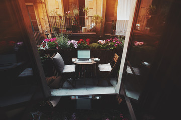 Small cozy beautiful workspace on balcony with laptop, mouse, smartphone, chairs and flowers around on sunny summer day, seeing through opened doors from dark interior, facade of another house behind