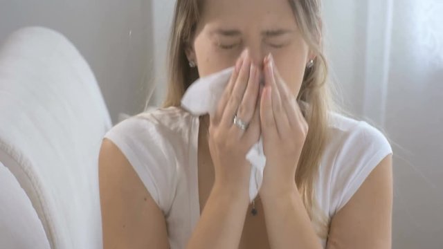 Closeup of young sick woman sitting on sofa at living room and sneezing in paper tissue. Footage shot in 4K