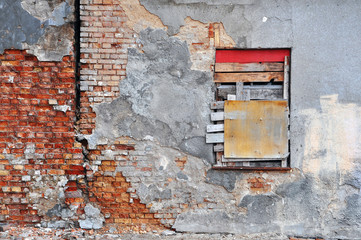 Gray wall of abandoned building with peeling plaster, red brick and the boarded-up window.