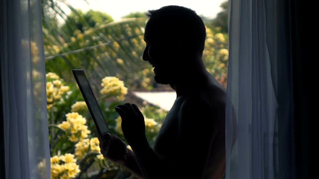 Silhouette of happy man browsing web on tablet by terrace window, super slow motion 240fps
