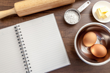Blank notebook with eggs, butter, wood roller and flour in measure cup on the wooden table