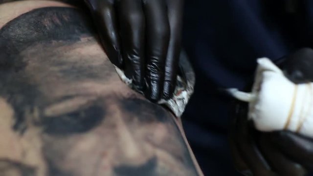 Tattoo master in black gloves, tattooing on the skin with black ink tattoo of his client in the salon.