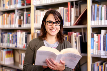 long brown hair girl with black glasses holding book and smiling. portrait of female student in a library with bookshelf on background