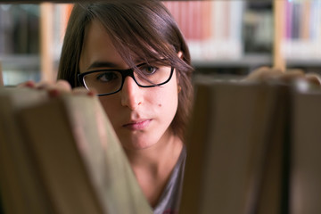 long brown hair girl with black glasses looking for a book that she needs. portrait of female student in a library with bookshelf on background