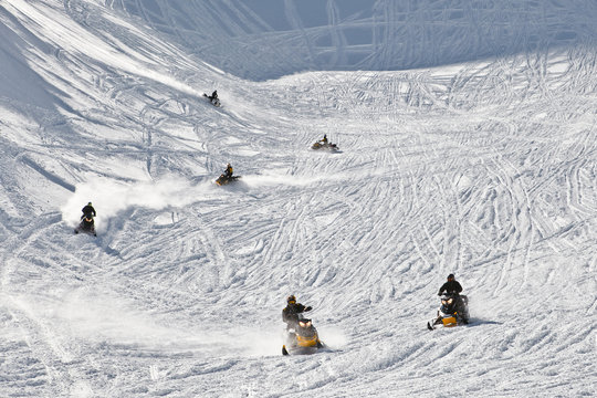 Men driving snowmobiles on snow covered mountainside tracks.