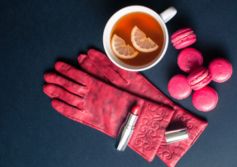 Cup of tea with french macaroon on dark background. Tea pot, cup of tea, lemon, red gloves, pink macaroon lying on black background.