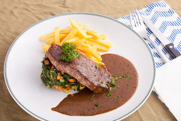 A dish of beef steak served with pepper brown sauce, spinach sauté and french fries in a decorated dish.