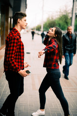 Two happy trendy teenage friends walking and dancing in the city, listening to the music with headphones, talking each other and smiling. Lifestyle, friendship and urban life concepts.