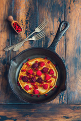 Homemade fried pancakes on a black cast iron skillet. Above are berries, raspberries, cranberries and blackberries. Wooden rustic brown background, vintage forks, top view.