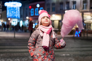 Little girl, iluminated with street lights, eating cotton candy. Christmas decoration is in the background