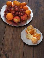 Plate with grapes, apples, grapes, clementine and an orange and a plate with peeled clementine on a wooden table.