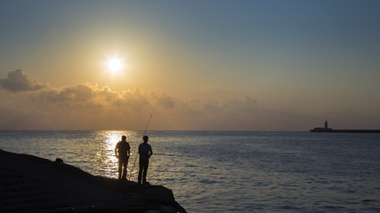 Fototapeta premium Valletta, Malta - Early in the morning two fishermen are waiting for a bite at Sliema Tigne point with Breakwater Lighthouse at background 