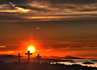 Three crosses, representing Jesus crucifixion, against a vibrant sunset sky, mountains and ocean.