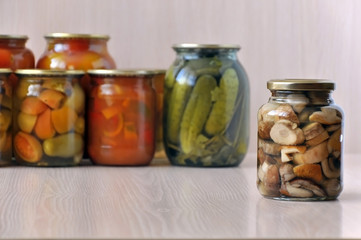 Glass jars with marinated mushrooms on a light wooden surface. Glass banks with vegetables on background. Close up.