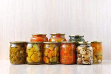Many inverted glass jars with canned pickled vegetables on wooden surface: peppers, tomatoes, cucumbers, mushrooms.