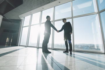 Two young businessmen are shaking hands with each other standing against panoramic windows.