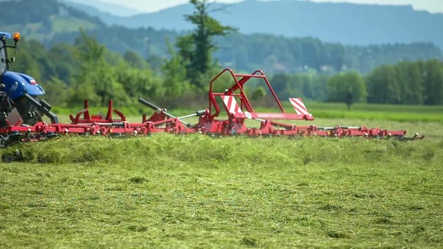 Red agricultural machinery is connected to a tractor and a farmer is out preparing hay. It is summer time.
