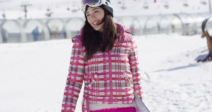 Cute and happy female asian snowboarder walking with her board on snowy ski slope. Wearing pink outfit and goggles. Enjoys her holidays.