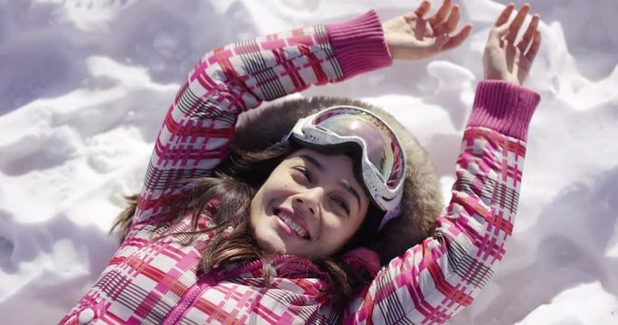 Half body portrait of happy young woman lying on snow with ski goggles and arms stetched out on ground