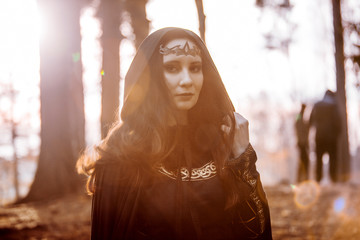 Young beautiful and mysterious woman in woods, in black cloak with hood, image of forest elf or...