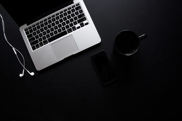 Laptop, mug, phone and earbuds on dark, black desk - obrazy, fototapety, plakaty