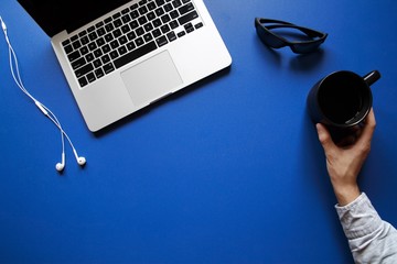 Hand placing mug next to laptop, sunglasses and earbuds on blue desk - 132445379