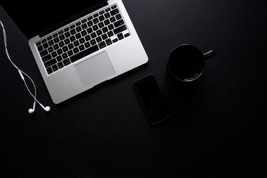 Laptop, mug, phone and earbuds on dark, black desk