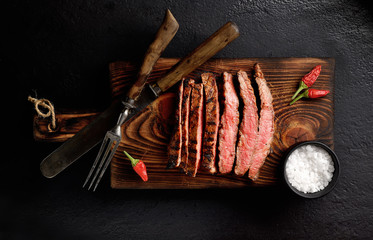 grilled beef on a board on a black background, knife, fork and salt