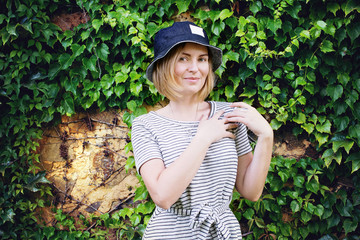 Pretty young woman in denim hat smiling and standing in front of natural wall with green leafs. - 132452751