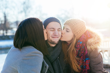 Two beautiful young women kissing handsome man standing between them. Winter outdoors fun.