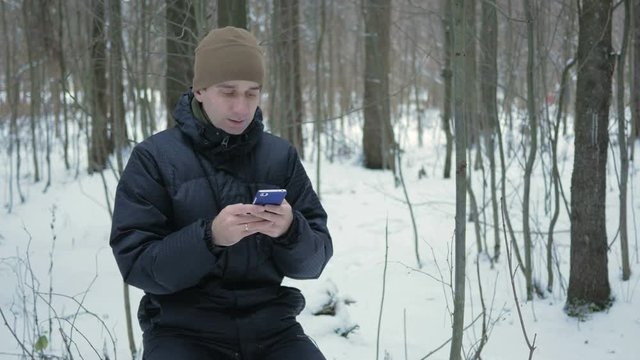 Young man typing a sms message on the phone in the winter snowy forest. He smiles and speaks the written.