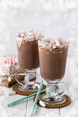Tasty cocoa drink with coloful marshmallows in two glasses on white wooden table. Chocolate beverage with paper straws.