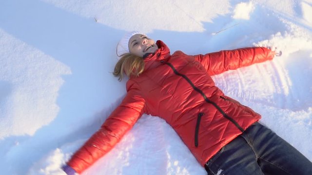 Beautiful young woman making snow angel in a winter park, having silly fun, smiling. Slow motion.
