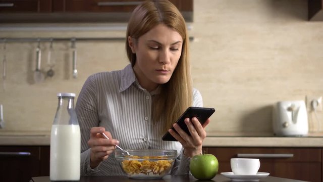 Beautiful young attractive businesswoman having cornflakes for breakfast and using her smartphone.