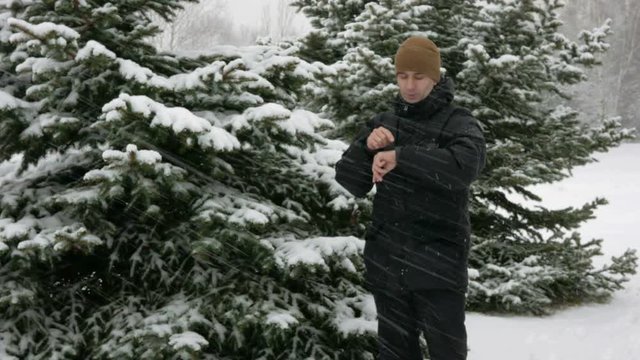 A man checks the messages on the smart watch in the winter snowy forest. Big snowfall. He dictates a voice response to the clock. A man dressed in a dark warm jacket.