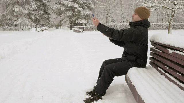 Man makes selfie phone in a snowy winter park. He smiles and changes the posture for a better photo. availability concept photo