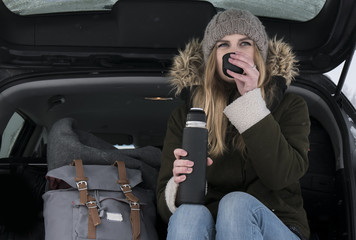 Young woman in a knitted shape sitting on trunk of a SUV and drinking hot tea from a thermos in...