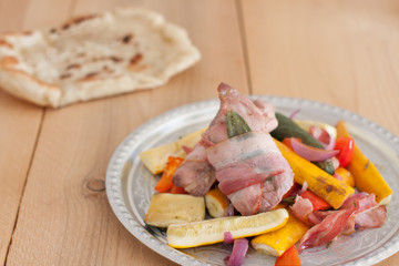 Pork tenderloin wrapped in bacon with colorful fresh roast vegetables - zucchini, bell pepper and onion and pita bread on a wooden board