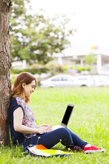 Beautiful student girl sitting on the grass and using laptop com